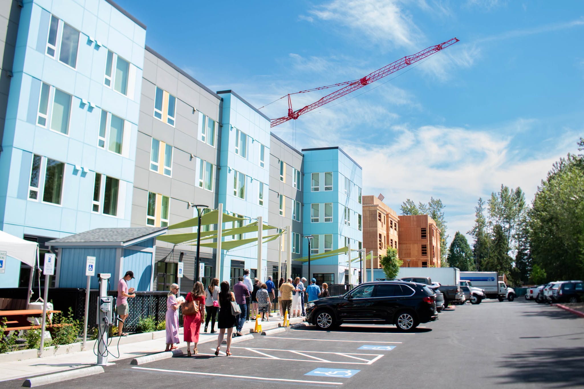 Group of people walking and standing in a parking lot beside a blue-gray modern building under a red construction crane in the sky.