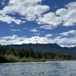 Snowy mountain rising above a dense forest along a calm lake under a blue sky with scattered clouds.