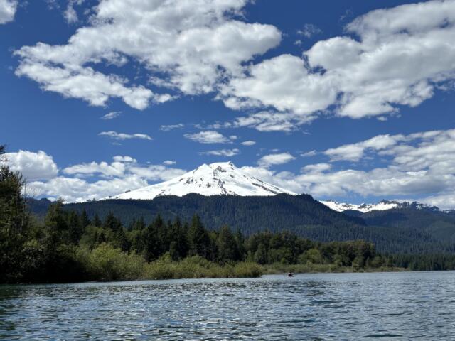Snowy mountain rising above a dense forest along a calm lake under a blue sky with scattered clouds.