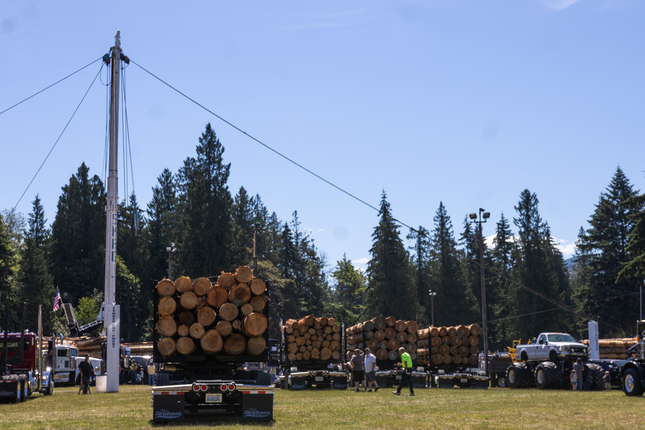 Flatbed trailers stacked with large logs at an outdoor logging event, with evergreen trees in the background and bystanders nearby.