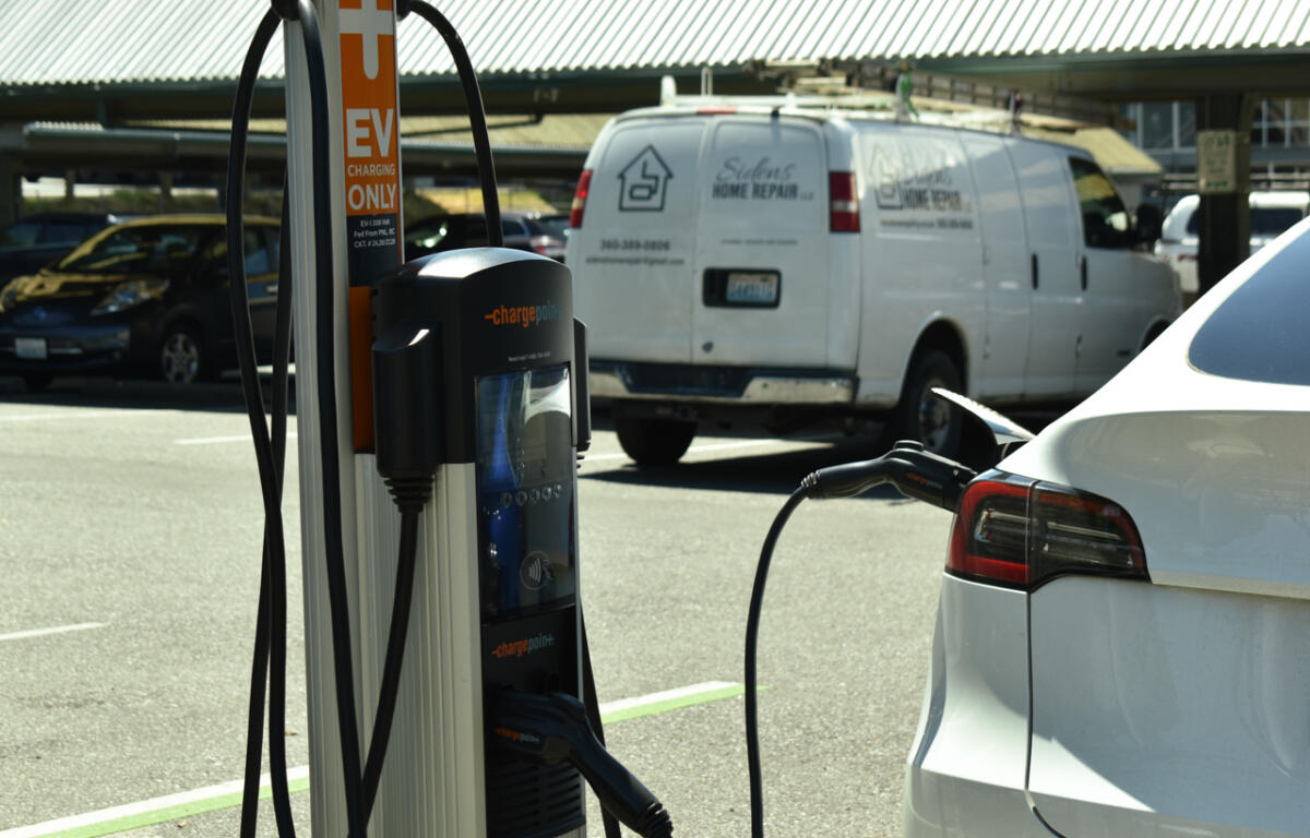 Electric vehicle charging station with a white car plugged in; orange and black ChargePoint unit in foreground, white van in background.