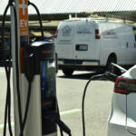 Electric vehicle charging station with a white car plugged in; orange and black ChargePoint unit in foreground, white van in background.