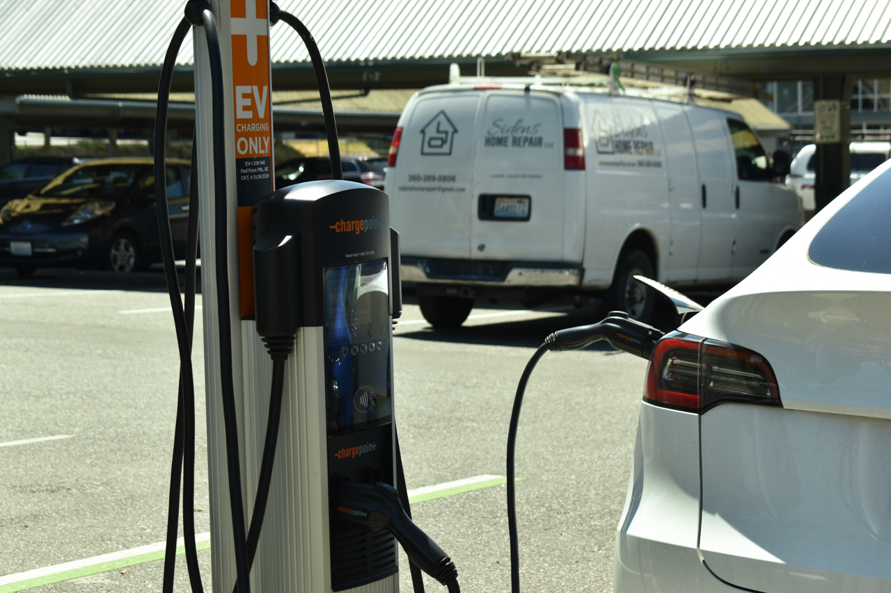 Electric vehicle charging station with a white car plugged in; orange and black ChargePoint unit in foreground, white van in background.