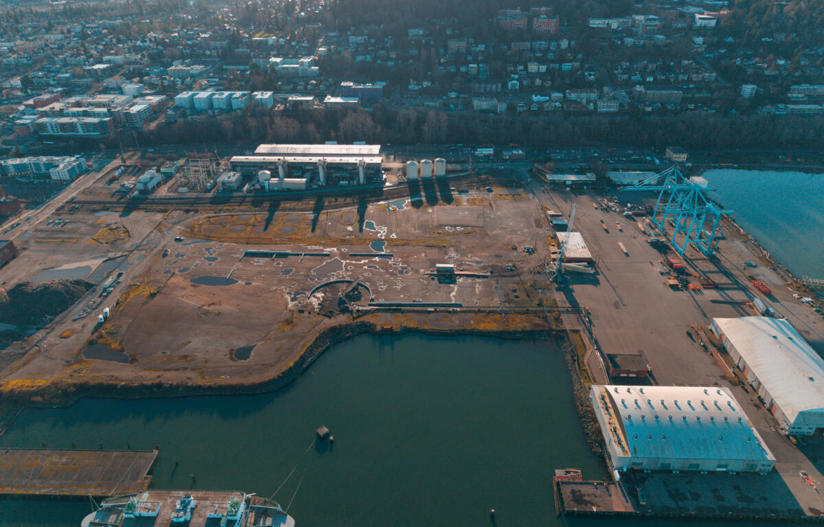 Aerial view of a harbor with a white passenger ship docked at the pier, cranes, and industrial buildings along the waterfront with a city beyond.