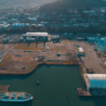 Aerial view of a harbor with a white passenger ship docked at the pier, cranes, and industrial buildings along the waterfront with a city beyond.