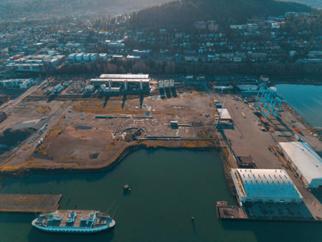 Aerial view of a harbor with a white passenger ship docked at the pier, cranes, and industrial buildings along the waterfront with a city beyond.