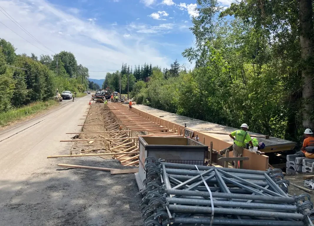 Road construction in a rural area with a long wooden formwork trench and workers installing materials under a blue sky.