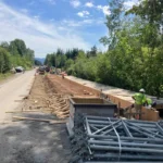 Road construction in a rural area with a long wooden formwork trench and workers installing materials under a blue sky.