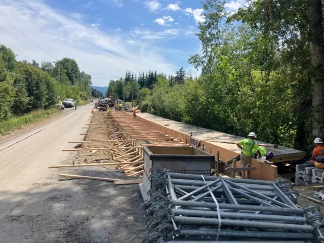 Road construction in a rural area with a long wooden formwork trench and workers installing materials under a blue sky.
