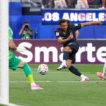 Soccer player in a dark kit kicks the ball toward goal as the green-clad goalkeeper dives, with a Los Angeles banner in the background.