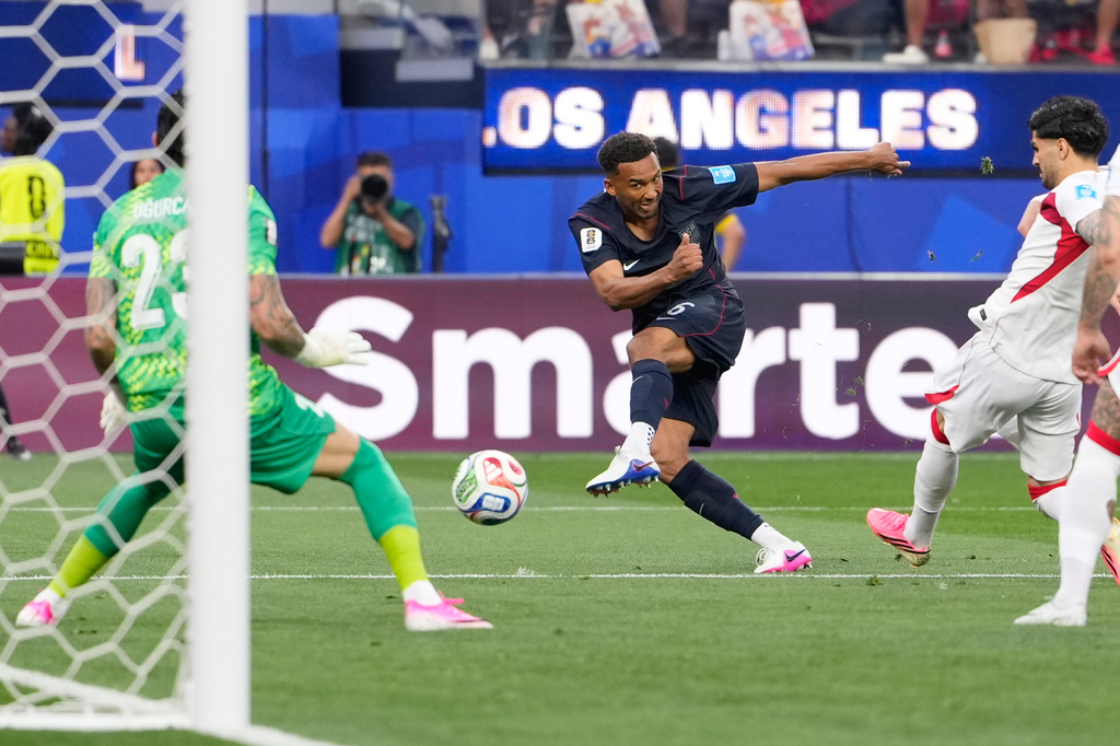 Soccer player in a dark kit kicks the ball toward goal as the green-clad goalkeeper dives, with a Los Angeles banner in the background.