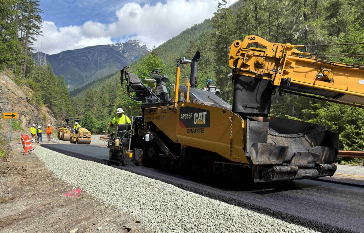 Construction crew and heavy asphalt paver laying gravel/road surface in a mountain forest; workers in safety vests nearby under blue sky
