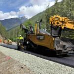 Construction crew and heavy asphalt paver laying gravel/road surface in a mountain forest; workers in safety vests nearby under blue sky