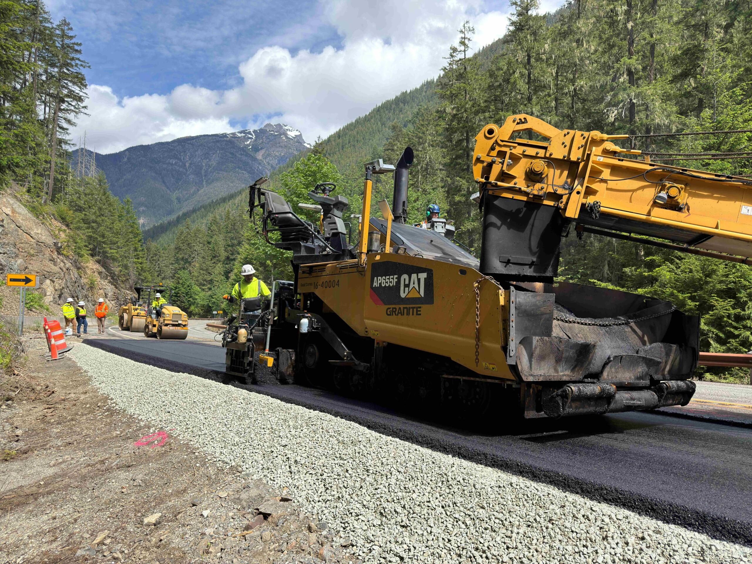 Construction crew and heavy asphalt paver laying gravel/road surface in a mountain forest; workers in safety vests nearby under blue sky