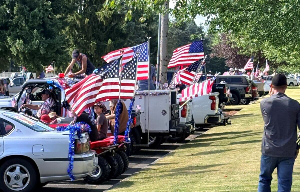 Patriotic parade scene with flag-draped trucks and American flags along a sunny street.