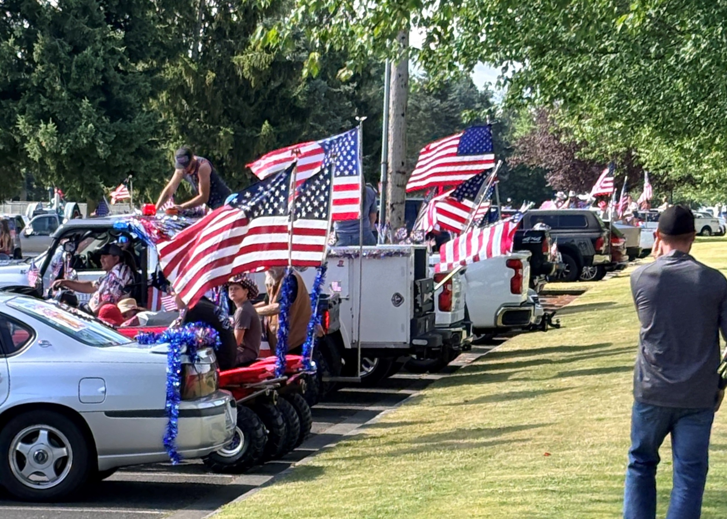 Patriotic parade scene with flag-draped trucks and American flags along a sunny street.