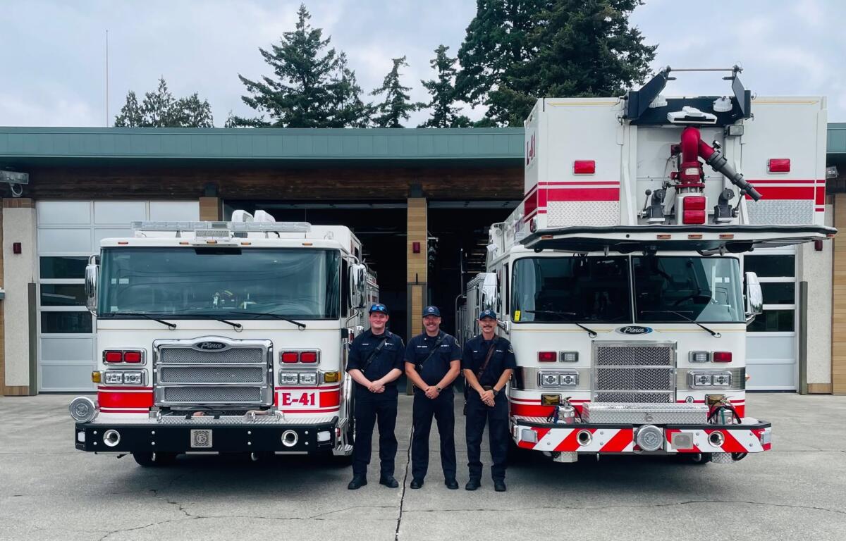 Four firefighters in dark uniforms stand in front of two red-and-white fire engines outside a fire station, smiling at the camera.