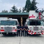 Four firefighters in dark uniforms stand in front of two red-and-white fire engines outside a fire station, smiling at the camera.