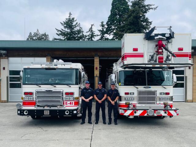 Four firefighters in dark uniforms stand in front of two red-and-white fire engines outside a fire station, smiling at the camera.