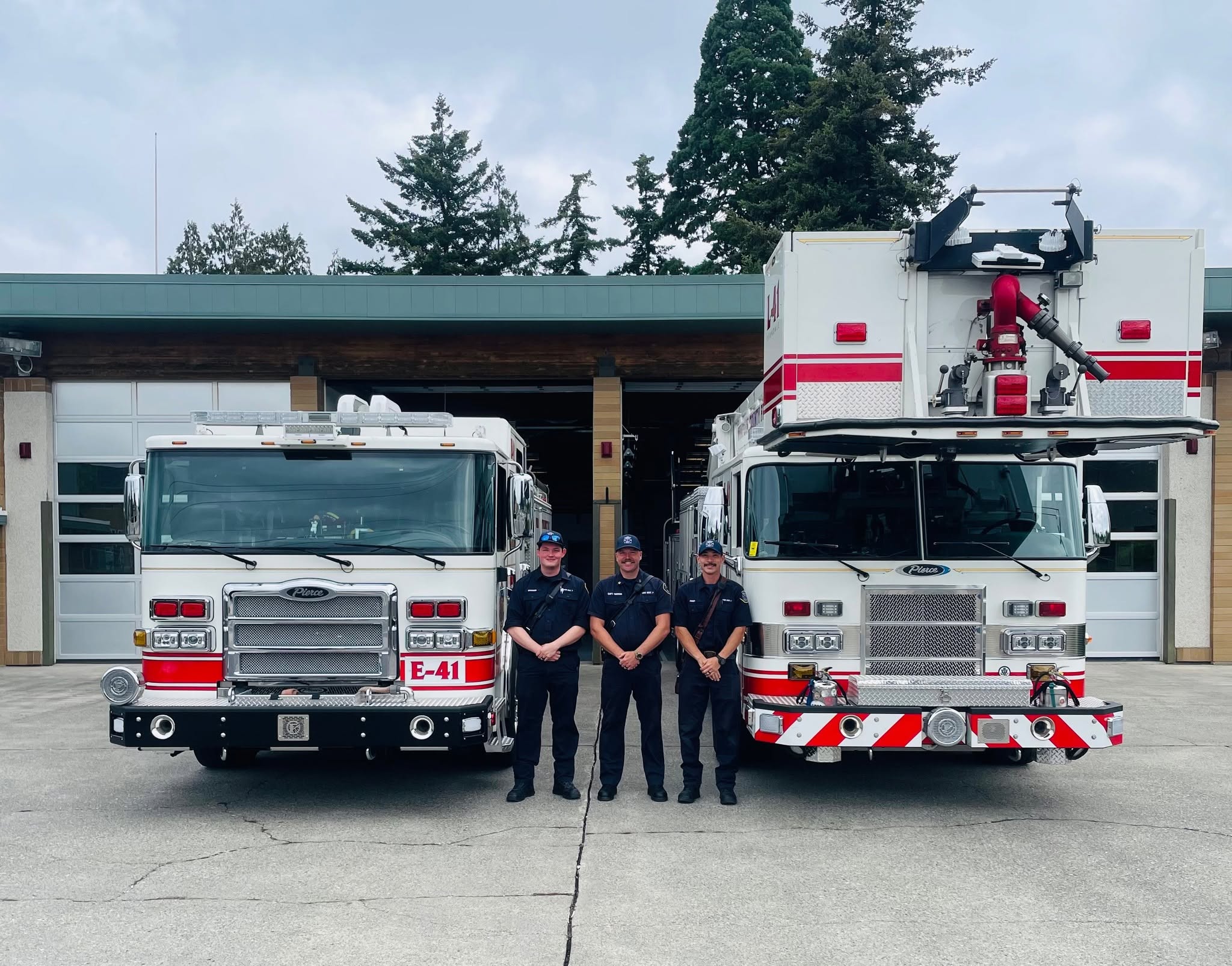 Four firefighters in dark uniforms stand in front of two red-and-white fire engines outside a fire station, smiling at the camera.