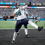 NFL player #98 in a white and blue jersey leaps in the end zone, holding the football in celebration with a packed stadium behind him.