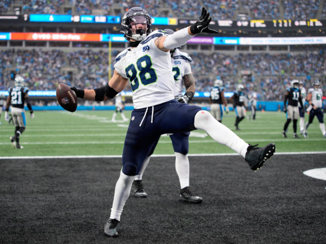 NFL player #98 in a white and blue jersey leaps in the end zone, holding the football in celebration with a packed stadium behind him.