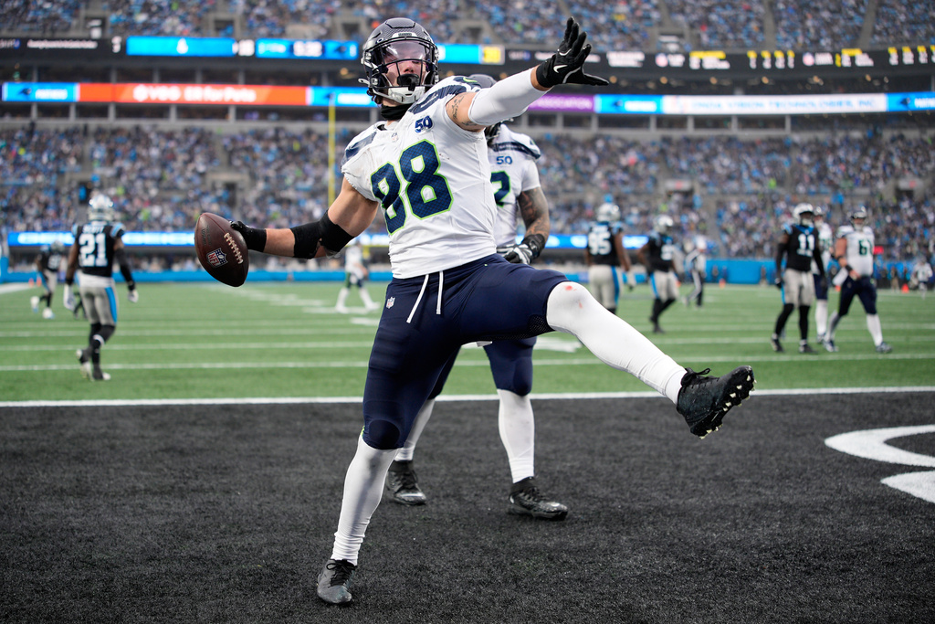 NFL player #98 in a white and blue jersey leaps in the end zone, holding the football in celebration with a packed stadium behind him.