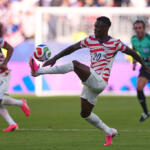USA soccer player number 20 stretches to kick the ball during a match, with a teammate and goalie in the background.