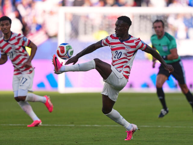 USA soccer player number 20 stretches to kick the ball during a match, with a teammate and goalie in the background.