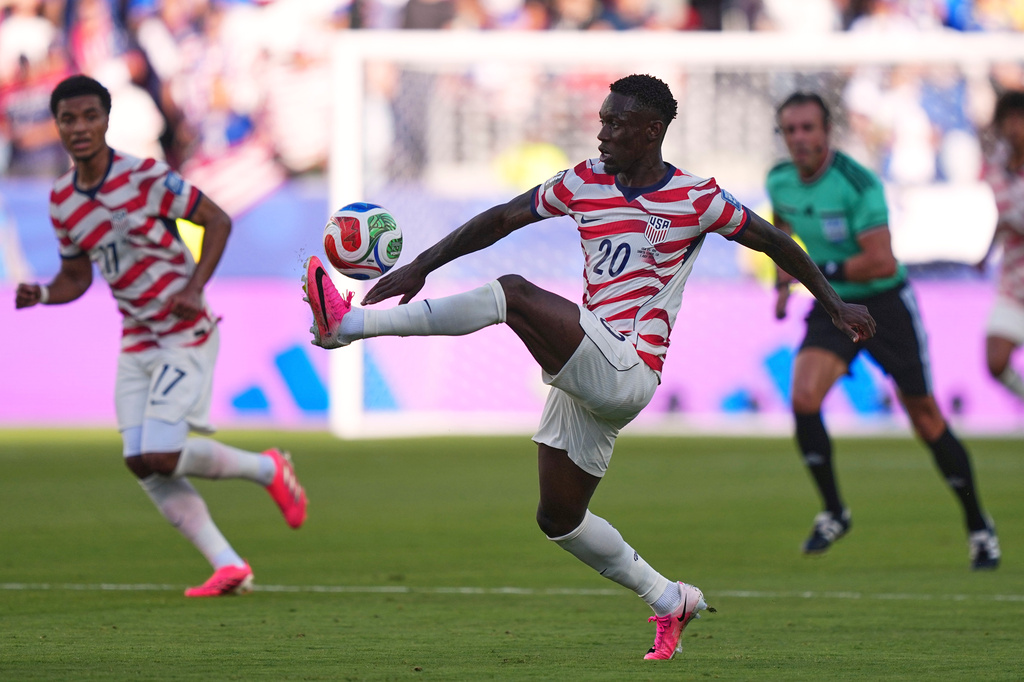 USA soccer player number 20 stretches to kick the ball during a match, with a teammate and goalie in the background.