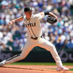 Right-handed pitcher in a beige Seattle jersey delivering a pitch on the mound with a crowded ballpark in the background}