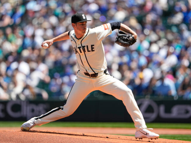 Right-handed pitcher in a beige Seattle jersey delivering a pitch on the mound with a crowded ballpark in the background}