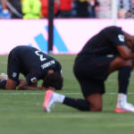 Two USA soccer players kneel and bow with heads down on a grass field during a match, showing distress or disappointment.