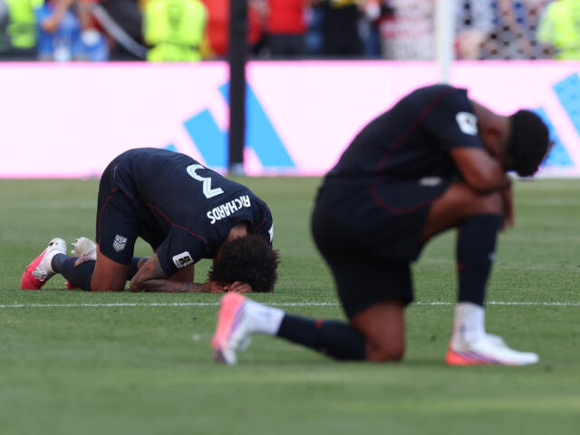 Two USA soccer players kneel and bow with heads down on a grass field during a match, showing distress or disappointment.