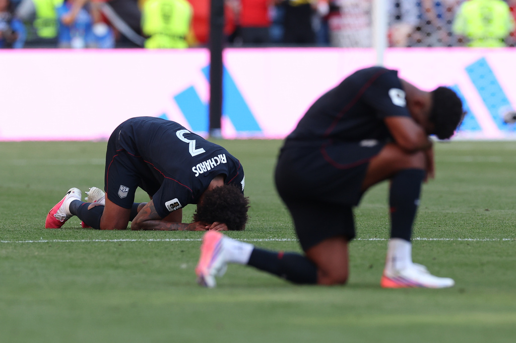 Two USA soccer players kneel and bow with heads down on a grass field during a match, showing distress or disappointment.