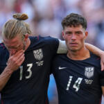 Two USA men’s soccer players with arms around each other after a match, wearing dark uniforms with numbers 13 and 14.