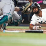 Baseball player in a white Miami uniform slides into home plate as the catcher reaches for a tag, dirt kicked up around the plate.
