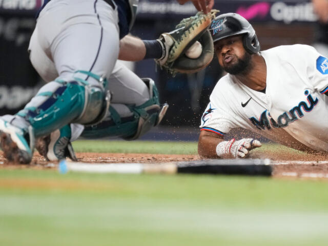 Baseball player in a white Miami uniform slides into home plate as the catcher reaches for a tag, dirt kicked up around the plate.