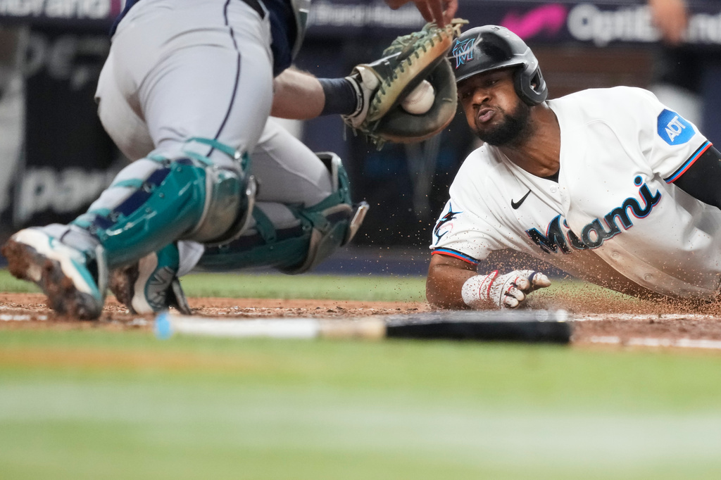 Baseball player in a white Miami uniform slides into home plate as the catcher reaches for a tag, dirt kicked up around the plate.