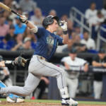 Baseball batter in Mariners uniform swings at a pitch with a catcher in blue gear behind him.