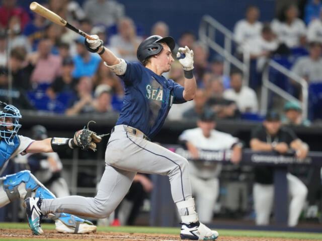 Baseball batter in Mariners uniform swings at a pitch with a catcher in blue gear behind him.