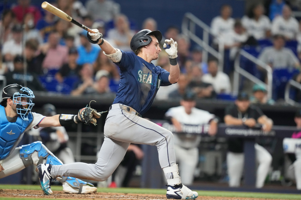 Baseball batter in Mariners uniform swings at a pitch with a catcher in blue gear behind him.