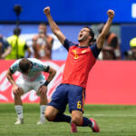 Soccer player in red Spain kit (number 5) raises fists in celebration on a green field, Coca-Cola boards behind him.
