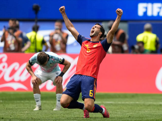 Soccer player in red Spain kit (number 5) raises fists in celebration on a green field, Coca-Cola boards behind him.