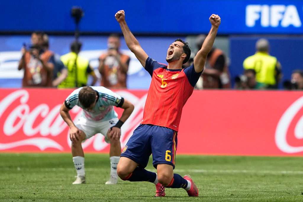 Soccer player in red Spain kit (number 5) raises fists in celebration on a green field, Coca-Cola boards behind him.