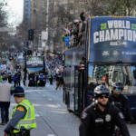 Blue double-decker parade bus with 'World Champions' sign, fans cheering on sidewalks and police in foreground.