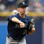 Seattle Mariners pitcher in a navy uniform throws a pitch during a game, with a glove on his left hand and a focused expression behind a blurred crowd.