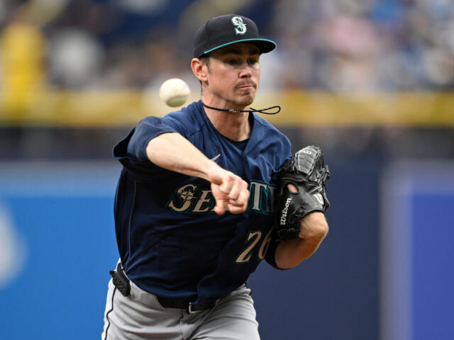 Seattle Mariners pitcher in a navy uniform throws a pitch during a game, with a glove on his left hand and a focused expression behind a blurred crowd.