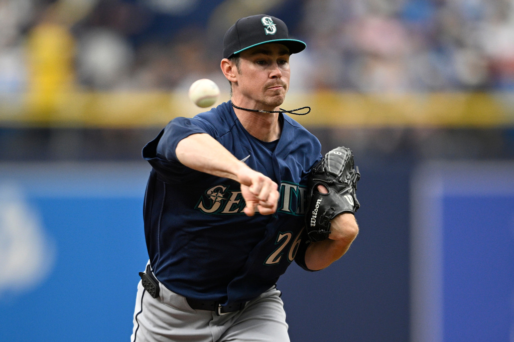 Seattle Mariners pitcher in a navy uniform throws a pitch during a game, with a glove on his left hand and a focused expression behind a blurred crowd.