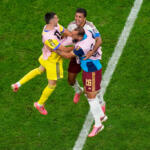 Three soccer players celebrate a goal, jumping and embracing on a green field near a white sideline line in mid-air.